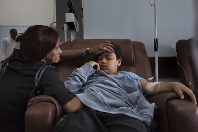 一个与登革热生病的孩子的照片，手臂上有静脉注射，一个女人轻轻地将手放在额头上